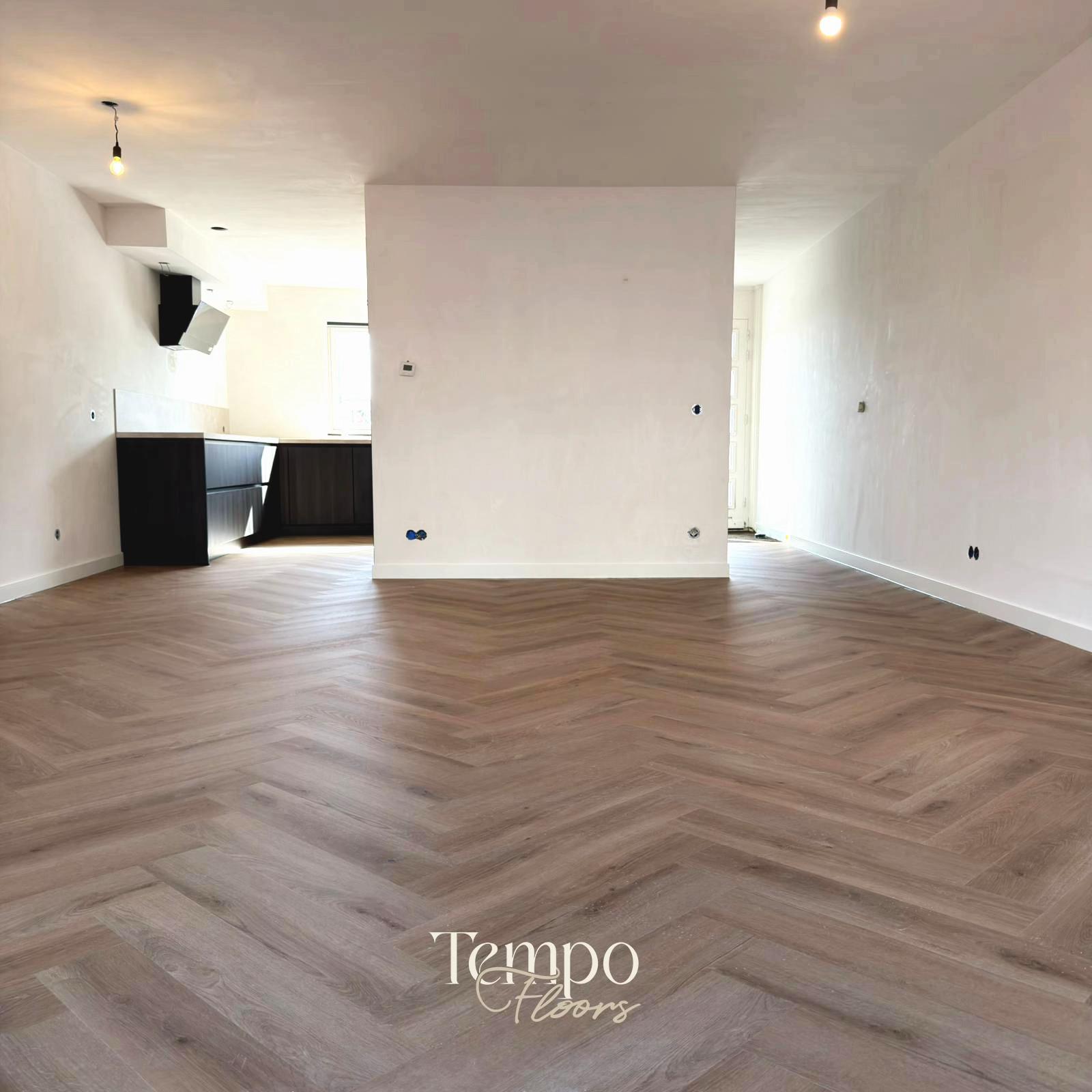 Een woonkamer met een lichte visgraatvloer, met de keuken op de achtergrond